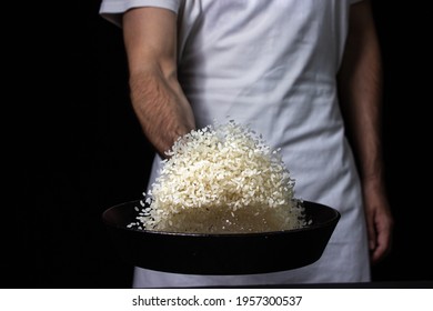 Toss The Rice In The Skillet. The Cook Is Throwing Rice On A Black Background. Cooking. Photo Of Food On A Dark Background