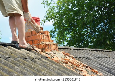 A total brick chimney rebuild. A building contractor on asbestos roof is removing bricks of a damaged chimney before a complete chimney rebuild.
