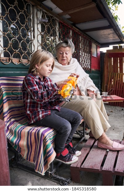 Total peace and\
understanding. Cheerful elderly lady giving advice to her male\
grandchildren about a plastic toy car while sitting next to each\
other near the country\
house