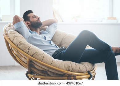Total relaxation. Handsome young man keeping eyes closed and holding hands behind head while sitting in big comfortable chair at home  