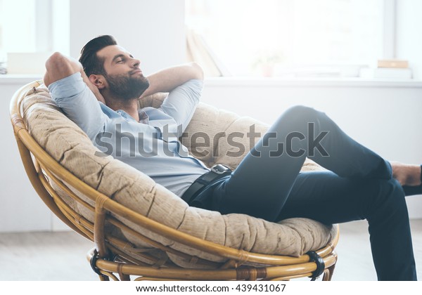 Total relaxation. Handsome young man keeping eyes closed\
and holding hands behind head while sitting in big comfortable\
chair at home  