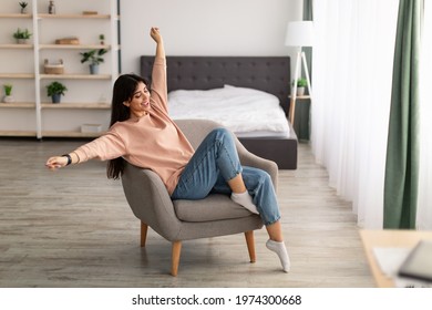 Total Relaxation And Satisfaction Concept. Portrait Of Beautiful Relaxed Woman Sitting On Comfortable Chair, Raising Arms And Stretching, Resting On Weekend Day In Living Room. Wellness, Wellbeing