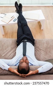Total relaxation. Top view of happy young African man in formalwear lying on the couch with his legs on a desk and hands behind head