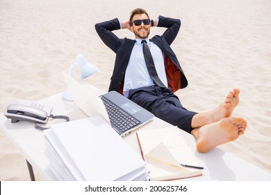 Total relaxation. Top view of relaxed young man in formalwear and sunglasses holding hands behind head and holding his feet on the table standing on sand