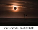 A total solar eclipse casts an eerie orange glow over a vast prairie landscape, silhouetting a lone windmill against the dramatic sky.