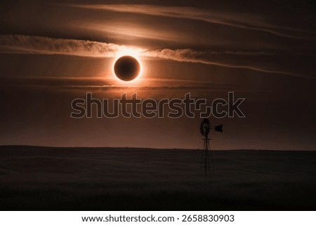 A total solar eclipse casts an eerie orange glow over a vast prairie landscape, silhouetting a lone windmill against the dramatic sky.