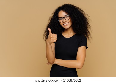 I Totally Agree. Good Looking Satisfied Afro American Woman Keeps Thumb Up, Dressed In Casual Black T Shirt, Has Crisp Hair, Wears Spectacles, Models Over Brown Background. Body Language Concept