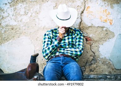 A Tough Cowboy Guy Sitting On A Wooden Fence. He Holds His Hat, Wears Jeans And Shirt. A Brown Saddle Is Next To Him. A Typical American Countryside Photo.