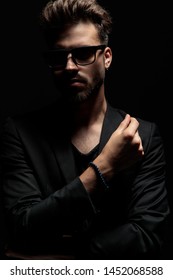 Tough dramatic man holding his hand crossed and looking forward while wearing a black jacket and sunglasses, standing on black studio background