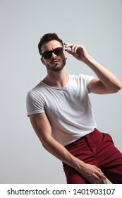 Tough Guy About To Take Off His Sunglasses While Wearing A White T-shirt And Red Pants, Sitting On White Studio Background