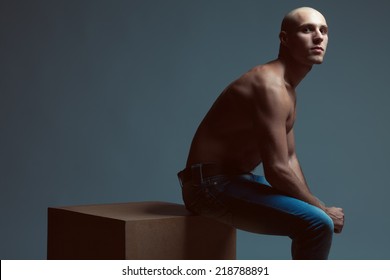 Tough Guy Concept. Handsome Bald Muscular Male Model In Blue Jeans With Perfect Muscular Body Posing Over Blue Background, Sitting On Wooden Cube. Vogue Style. Copy-space. Fashion Studio Portrait