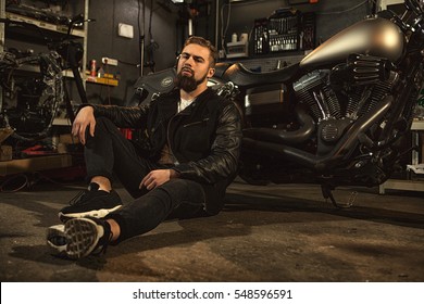 Tough Guy. Low Angle Shot Of A Handsome Bearded Young Biker Guy Sitting On The Floor Near His Chopper Motorcycle Relaxing At The Repair Shop Copyspace Lifestyle Biker Workshop Concept