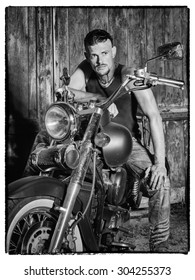 Tough Guy With Sparrow Beard, Undercut And Blue Jeans Sitting On A Chopper Bike In Front Of  A Green Weathered Barn Door