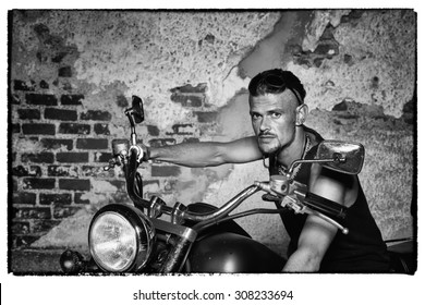 Tough Guy With Sparrow Beard, Undercut And Blue Jeans Sitting On A Chopper Bike In Front Of  A Weathered Brick Wall