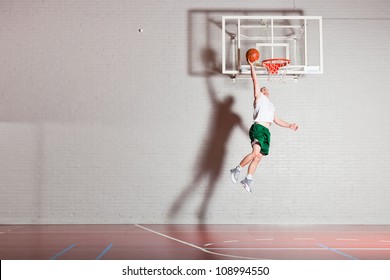 Tough Healthy Young Man Playing Basketball In Gym Indoor. Wearing White Shirt And Green Shorts.