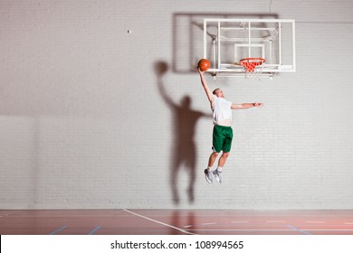 Tough Healthy Young Man Playing Basketball In Gym Indoor. Wearing White Shirt And Green Shorts.