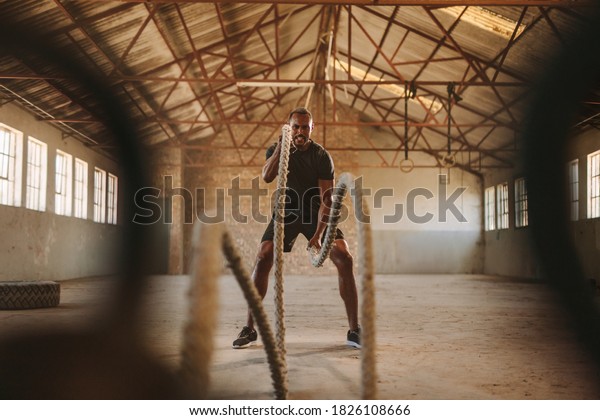 Tough man working out in training gym made\
inside old factory. Fitness man exercising with battle rope in\
abandoned warehouse.