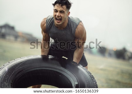 Tough young male athlete doing a tire flip exercise in the rain. Muscular man doing cross training outdoors on field.
