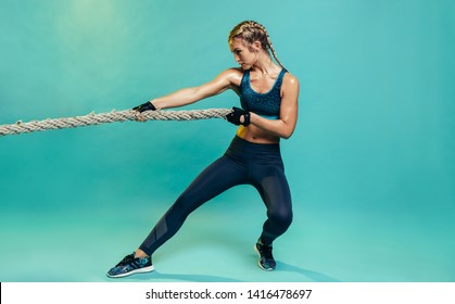Tough young woman exercising with battling rope in studio. Healthy sports woman working out with battle rope over blue background.