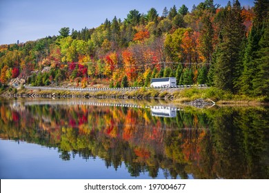 Tour bus driving though fall forest with colorful autumn leaves reflecting in lake. Highway 60 at Lake of Two Rivers, Algonquin Park, Ontario, Canada.