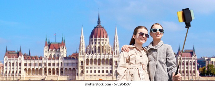 Tourism, Childhood And Travel Concept - Happy Girls Taking Picture With Smartphone On Selfie Stick Over House Of Parliament In Budapest