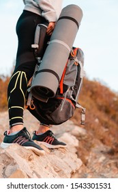 Tourism And Sports Recreation. A Woman Holds A Tourist Backpack And A Mat. Close-up Of Legs And Sports Equipment. Vertical Orientation