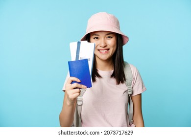 Tourism and tourists concept. Smiling happy asian girl traveller, wearing panama hat, showing passport with flight tickets, booking trip, blue background