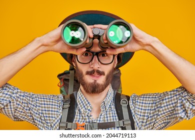 Tourism And Travel, Adventure. Funny Excited Young Man With Backpack And Tourist Hat Looking Through Binoculars. Studio Portrait In A Yellow Background.