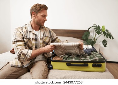 Tourism and travelling concept. Portrait of handsome young man going on holiday, packing his suitcase, checking luggage, sitting on bed and looking at packed things. - Powered by Shutterstock