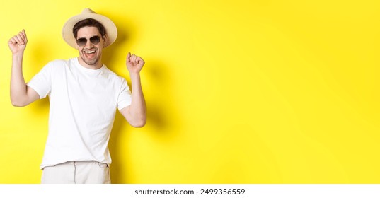 Tourism, travelling and holidays concept. Happy caucasian guy dancing and having fun on vacation, wearing sunglasses with straw hat, standing against yellow background. - Powered by Shutterstock