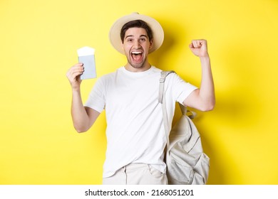 Tourism and vacation. Man feeling happy about summer trip, holding passport with plane tickets and backpack, raising hands up in celebration gesture, yellow background