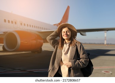 tourism, vacation and people concept - happy smiling young woman wearing hat with travel bag pack standing by the airplane and ready to departure. - Shutterstock ID 2172808657