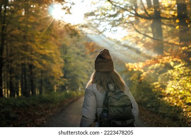 Tourist with backpack and knit hat hiking in autumn forest. Woman enjoying sunrise during hike in woodland. Outdoors activity in fall season - Powered by Shutterstock