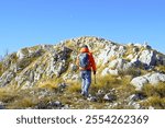 A tourist in a bright orange jacket, photographed from behind, walks along the Dobroštica ridge in the Orjen Nature Park, Montenegro. A man with hiking poles walks in the mountains above Herceg Novi.