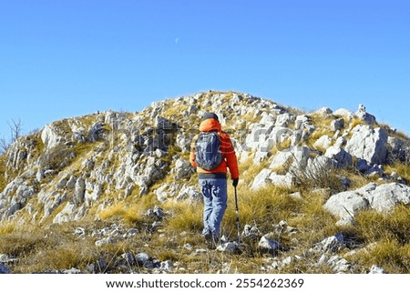 A tourist in a bright orange jacket, photographed from behind, walks along the Dobroštica ridge in the Orjen Nature Park, Montenegro. A man with hiking poles walks in the mountains above Herceg Novi.
