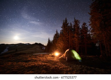 Tourist Camping Near Forest In The Night. Illuminated Tent And Campfire Under Beautiful Night Sky Full Of Stars And The Moon