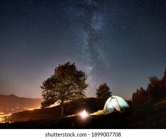 Tourist Camping in der Nähe des Waldes in der Sommernacht. Zelt und Lagerfeuer unter erstaunlichem Nachthimmel voller Sterne und Milchstraße. Auf dem Hintergrund schöner Sternenhimmel, Berge und helle Stadt
