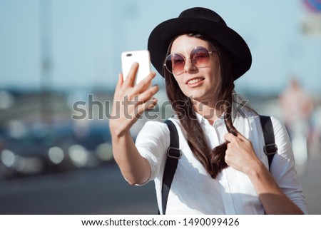Tourist in the city takes a photo on your smartphone. Young woman in black hat and white shirt. Funny hipster girl takes selfie on phone