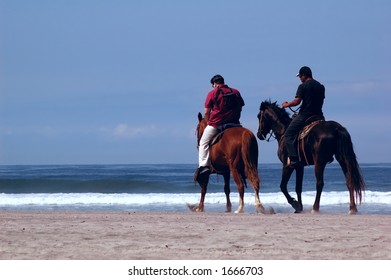 A tourist and a cowboy ride horses down an ocean beach