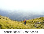 A tourist descends a steep mountainside covered with bright yellow grass toward the Сarine katun. The man walks along a path against the backdrop of a mountain valley with low clouds hanging above.