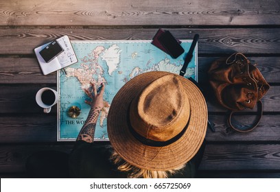 Tourist Exploring The World Map Sitting On Wooden Floor With Compass And Other Travel Accessories. Young Woman Wearing Brown Hat Looking At The World Map.