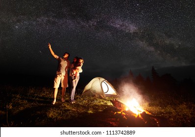 Tourist Family Camping In Mountains At Night. Mother Holding In Arms Small Daughter, Father Pointing At Bright Stars In Dark Sky And Milky Way In Front Of Illuminated Tent And Burning Bonfire