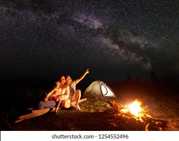 Tourist Family Camping In Mountains At Night, Sitting On Log Near Illuminated Tent And Burning Bonfire. Mother Holding In Arms Small Daughter, Father Pointing At Bright Stars In Dark Sky And Milky Way