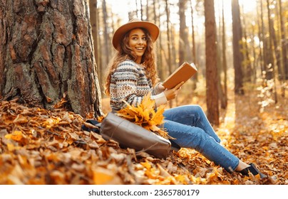 A tourist female in a hat sits in the autumn forest on yellow leaves and reads a book. Beautiful woman enjoying sunny autumn weather. - Shutterstock ID 2365780179