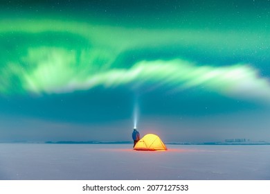 Tourist mit Taschenlampe nahe gelbem Zelt von innen beleuchtet vor dem Hintergrund von unglaublichem Sternenhimmel mit Aurora borealis. Erstaunliche Nachtlandschaft. Nordlichter im Winterfeld