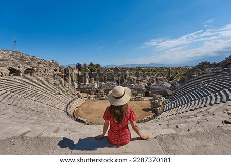 Tourist girl with hat and red dress posing, sitting and watching panorama of amphitheatre in ancient city of Side, Manavgat, Antalya, Turkey