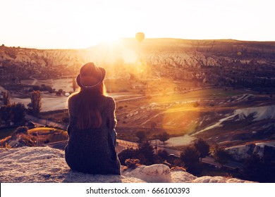 A tourist girl in a hat sits on a mountain and looks at the sunrise and balloons in Cappadocia. Tourism, sightseeing, Turkey.