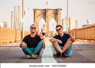 Tourist handsome models wearing sunglasses posing for a photo and making peace sign on a Brooklyn Bridge with unrecognizable blurry people in background during sunny summer day In New York City, USA - Powered by Shutterstock