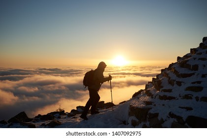 Tourist Hiker With Backpack Climbing Up On Rocky Mountain Steep Slope Covered With First Snow On Background Of Foggy Valley Filled With White Puffy Clouds, Raising Sun And Bright Blue Sky At Dawn.