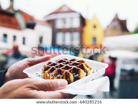 Tourist holds in hand popular street food - Belgium tasty waffle with chocolate sauce on the background of city tourist streets of Bruges, Belgium, Europe. Traditional Belgian dessert, pastry.